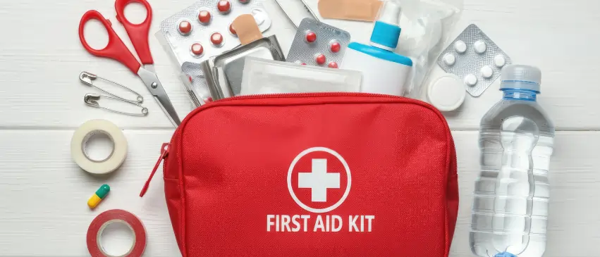 Red workplace first aid kit surrounded by medical supplies including bandages, pills, scissors, tape, antiseptic solution, and a water bottle on a white surface.