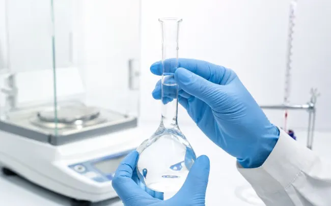 A laboratory technician wearing blue gloves carefully checks the liquid meniscus in a clear volumetric flask, with an analytical balance and laboratory equipment blurred in the background.